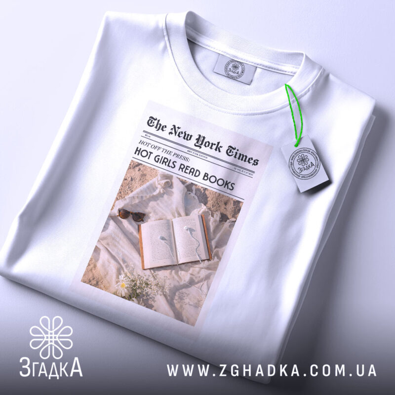 Футболка дівчата читають книги, біла, з верхнього ракурсу, складена на поверхні, фон світлий, принт у стилі газети з текстом 'HOT GIRLS READ BOOKS' та зображенням книги з навушниками.