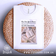 Футболка дівчата читають книги, біла, з верхнього ракурсу, складена на плетеній кошику, фон натуральний, принт у стилі газети з текстом 'HOT GIRLS READ BOOKS' та зображенням книги з навушниками.