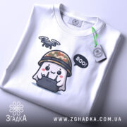 Футболка Дрон під контролем, біла, складена на столі, фон світло-сірий, принт з веселим привидом у камуфляжному шоломі, який тримає пульт управління з дроном, текст 'BOO!' у бульбашці, на комірці видно етикетку.