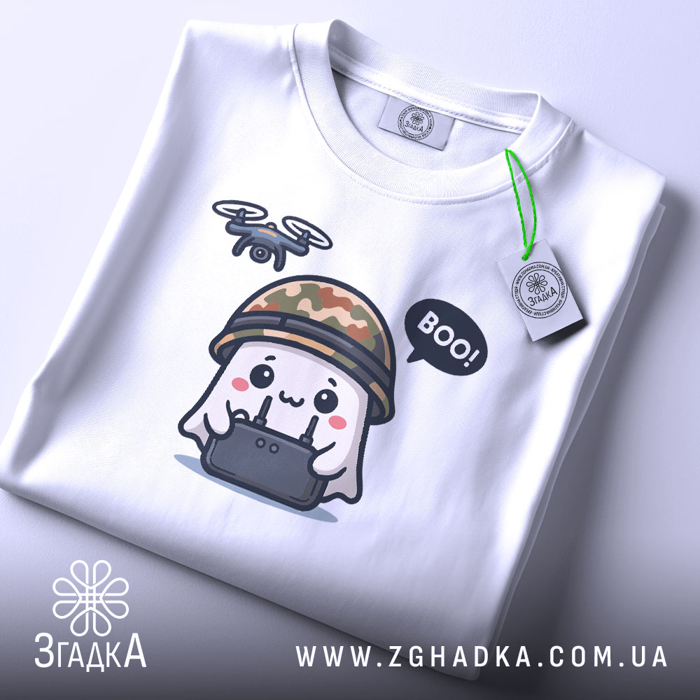 Футболка Дрон під контролем, біла, складена на столі, фон світло-сірий, принт з веселим привидом у камуфляжному шоломі, який тримає пульт управління з дроном, текст 'BOO!' у бульбашці, на комірці видно етикетку.