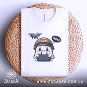 Футболка Дрон під контролем, біла, складена на кошику з плетеного матеріалу, фон світло-сірий, принт з веселим привидом у камуфляжному шоломі, який тримає пульт управління з дроном, текст 'BOO!' у бульбашці.