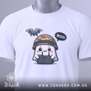 Футболка Дрон під контролем, біла, складена на столі, фон світло-сірий, принт з веселим привидом у камуфляжному шоломі, який тримає пульт управління з дроном, текст 'BOO!' у бульбашці, на комірці видно етикетку.