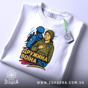 Футболка дружина воїна ЗСУ, біла, складена на поверхні, фон світло-сірий, принт з жінкою в капюшоні та військовим, кольори принта: жовтий, синій, зелений, з біркою на шнурку.