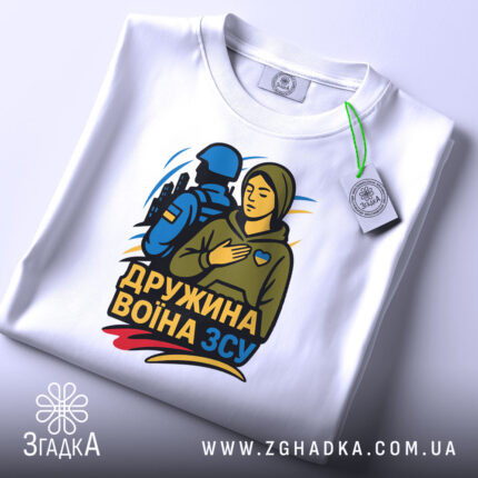 Футболка дружина воїна ЗСУ, біла, складена на поверхні, фон світло-сірий, принт з жінкою в капюшоні та військовим, кольори принта: жовтий, синій, зелений, з біркою на шнурку.