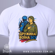 Футболка дружина воїна ЗСУ, біла, складена на поверхні, фон світло-сірий, принт з жінкою в капюшоні та військовим, кольори принта: жовтий, синій, зелений, з біркою на шнурку.