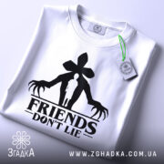 Футболка друзі не брешуть, біла, зображена зверху, складена на поверхні, фон світло-сірий, принт чорного кольору з силуетом людини та великими руками, на футболці є етикетка з зеленим шнурком.