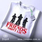 Футболка Друзі не брешуть білого кольору, складена на столі, фон світло-сірий, принт з трьома фігурами дітей на велосипедах у чорному кольорі, текст 'FRIENDS DON'T LIE' червоного кольору під принтом, на футболці є етикетка з зеленим шнурком.