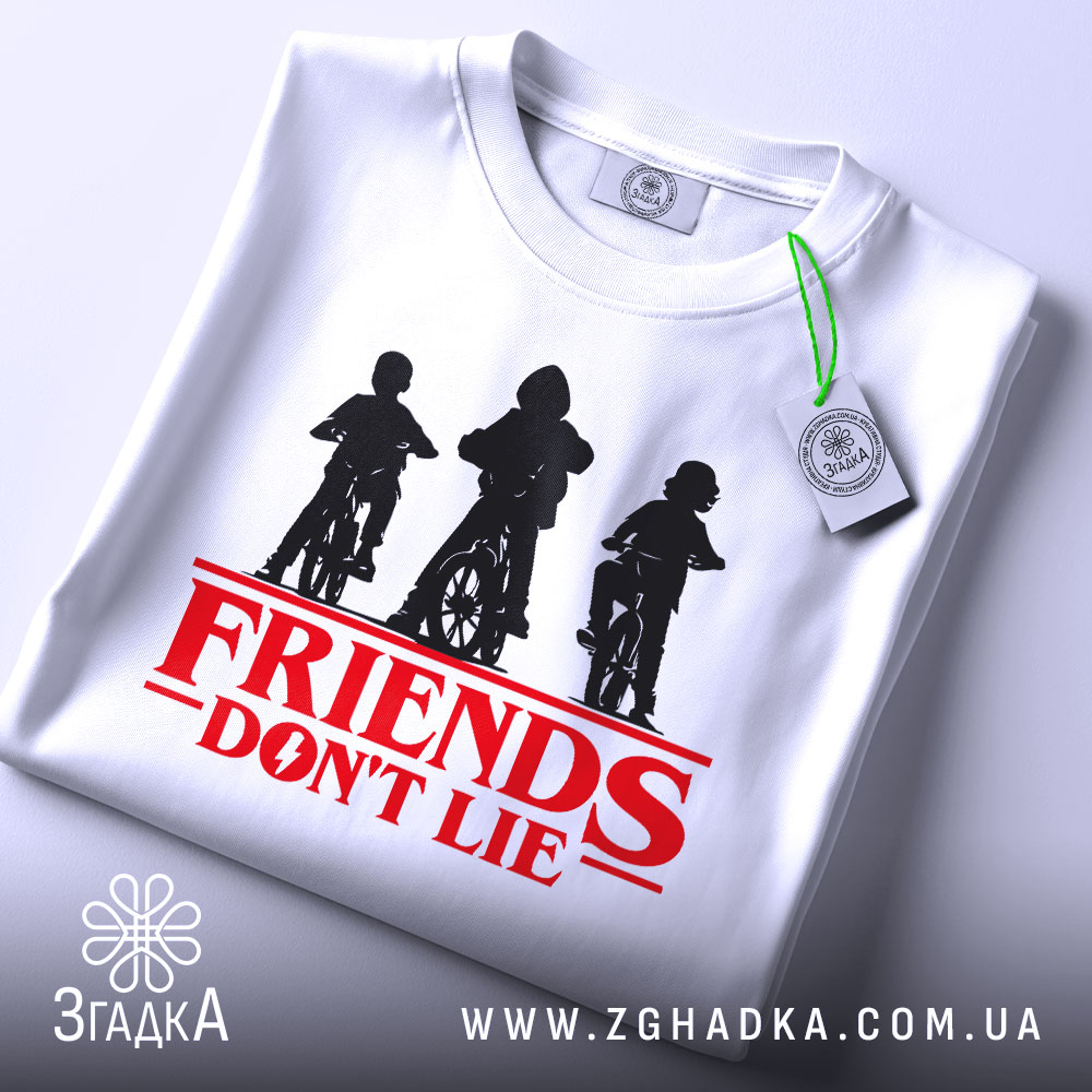 Футболка Друзі не брешуть білого кольору, складена на столі, фон світло-сірий, принт з трьома фігурами дітей на велосипедах у чорному кольорі, текст 'FRIENDS DON'T LIE' червоного кольору під принтом, на футболці є етикетка з зеленим шнурком.