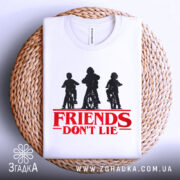 Футболка Друзі не брешуть білого кольору, складена на плетеній кошику, фон світло-сірий, принт з трьома фігурами дітей на велосипедах у чорному кольорі, текст 'FRIENDS DON'T LIE' червоного кольору під принтом.