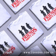 Футболка Друзі не брешуть білого кольору, зображена на манекені з правого боку, фон світло-сірий, принт з трьома фігурами дітей на велосипедах у чорному кольорі, текст 'FRIENDS DON'T LIE' червоного кольору під принтом.