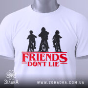 Футболка Друзі не брешуть білого кольору, складена на столі, фон світло-сірий, принт з трьома фігурами дітей на велосипедах у чорному кольорі, текст 'FRIENDS DON'T LIE' червоного кольору під принтом, на футболці є етикетка з зеленим шнурком.