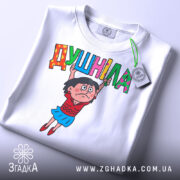 Футболка душніла білого кольору, складена на поверхні, з принтом, що зображає персонажа, який тримає напис "Душніла". На футболці є етикетка, фон світлий.