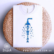 Футболка душніла білого кольору, складена на плетеній кошику, фон білий, принт з душем та написом "Душніла" синього кольору, розташований по центру.