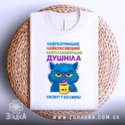 Футболка душніла експерт, біла футболка з принтом, зображення кота з блакитною шерстю, що тримає жовту чашку, текст на футболці кольоровий: синій, зелений, фіолетовий, червоний, футболка лежить на плетеній основі.