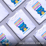 Футболка душніла експерт, біла футболка з принтом, зображення кота з блакитною шерстю, що тримає жовту чашку, текст на футболці кольоровий: синій, зелений, фіолетовий, червоний, фон світло-сірий.