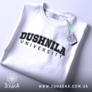 Футболка душніла білого кольору, складена на столі, фон світло-сірий, принт чорного кольору з написом 'DUSHNILA UNIVERSITY', на комірці видно етикетку.