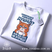 Футболка душніла білого кольору, зображена з верхнього ракурсу, фон світло-сірий, принт з котом, що тримає чашку, текст на принті синього кольору, на футболці є етикетка з зеленим шнурком.