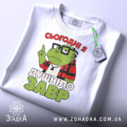 Футболка душнілозавр білого кольору, складена на поверхні, фон світло-сірий, принт з зеленим персонажем у червоній клітинці та написом 'СЬОГОДНІ Я ДУШНІЛО ЗАВР', з етикеткою на зеленому шнурку.