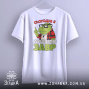 Футболка душнілозавр білого кольору, підвішена на вішалці, фон світло-сірий, принт з зеленим персонажем у червоній клітинці та написом 'СЬОГОДНІ Я ДУШНІЛО ЗАВР'.