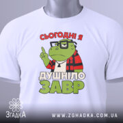 Футболка душнілозавр білого кольору, складена на поверхні, фон світло-сірий, принт з зеленим персонажем у червоній клітинці та написом 'СЬОГОДНІ Я ДУШНІЛО ЗАВР', з етикеткою на зеленому шнурку.