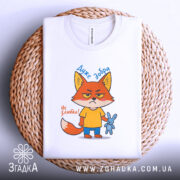 Футболка Дуже добра, білого кольору, зображена зверху на плетеній кошику, принт з мультяшним лисом у жовтій футболці, який тримає синю іграшку-кролика, текст "Дуже добра" та "Не зліть!", фон світлий.