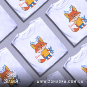 Футболка Дуже добра, білого кольору, зображена в анфас на манекені, фон світло-сірий, принт з мультяшним лисом у жовтій футболці, який тримає синю іграшку-кролика, текст зверху "Дуже добра", а знизу "Не зліть!".