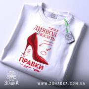 Футболка диявол правки вносить, біла, складена на поверхні, фон світлий, принт червоного кольору з малюнком черевика на високих підборах та текстом, з зеленим ярликом.