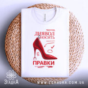 Футболка диявол правки вносить, біла, складена на плетеній кошику, фон світлий, принт червоного кольору з малюнком черевика на високих підборах та текстом.