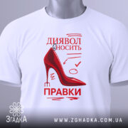 Футболка диявол правки вносить, біла, складена на поверхні, фон світлий, принт червоного кольору з малюнком черевика на високих підборах та текстом, з зеленим ярликом.