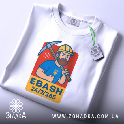 Футболка EBASH білого кольору, зображена зверху, на ній видно принт з чоловіком у жовтій касці з киркою, на синій футболці, з написом 'EBASH 24/7/365', на футболці є етикетка з зеленим шнурком.