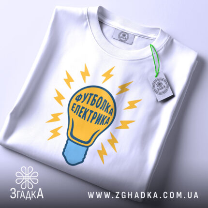 Футболка електрика 220 вольт, біла, складена на столі, фон світло-сірий, принт у вигляді лампочки з жовтими блискавками та написом 'Футболка електрика', на ярлику зелена стрічка.