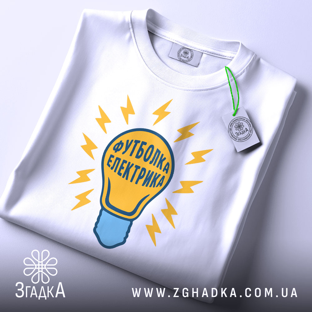 Футболка електрика 220 вольт, біла, складена на столі, фон світло-сірий, принт у вигляді лампочки з жовтими блискавками та написом 'Футболка електрика', на ярлику зелена стрічка.