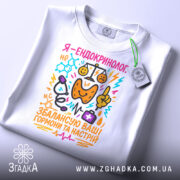 Футболка ендокринолог, біла, складена на столі, фон світло-сірий, принт кольоровий, текст "Я - ендокринолог. Збалансую ваші гормони та настрій" з ілюстраціями медичних інструментів.