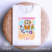 Футболка ендокринолог, біла, складена на плетеній кошику, фон світло-сірий, принт кольоровий, текст "Я - ендокринолог. Збалансую ваші гормони та настрій" з малюнками медичних символів.