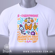 Футболка ендокринолог, біла, складена на столі, фон світло-сірий, принт кольоровий, текст "Я - ендокринолог. Збалансую ваші гормони та настрій" з ілюстраціями медичних інструментів.
