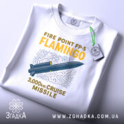 Футболка фламіго білого кольору, складена на поверхні, ракурс зверху, фон світло-сірий, принт з малюнком ракети та текстом "FIRE POINT FP-5 FLAMINGO 3,000km CRUISE MISSILE" у жовтому та синьому кольорах, з етикеткою зеленого кольору.