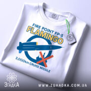 Футболка фламінго крилата ракета білого кольору, зображена на столі з верхнього ракурсу, фон світло-сірий, принт містить синю ракету та червоний хрест, текст на футболці жовтого кольору, прикріплена етикетка зеленого кольору.