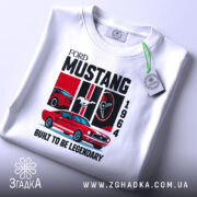 Футболка Ford Mustang класика, біла, складена на поверхні, фон світло-сірий, принт з червоним автомобілем Ford Mustang 1964, логотипом Ford, написом 'BUILT TO BE LEGENDARY', з етикеткою на рукаві.