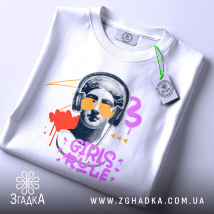 Футболка Girls Rule Art білого кольору, складена на поверхні, зображена з верхнього ракурсу, фон світло-сірий, принт з графічним зображенням статуї з навушниками та сонцезахисними окулярами, кольори принта: червоний, фіолетовий, жовтий, чорний, на футболці є етикетка.