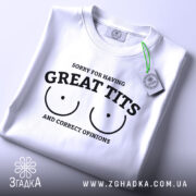 Футболка great tits білого кольору, складена на столі, ракурс зверху, фон світло-сірий, принт чорного кольору з текстом 'SORRY FOR HAVING GREAT TITS AND CORRECT OPINIONS' і малюнком двох грудей, на футболці є зелений ярлик.