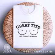 Футболка great tits білого кольору, складена на плетеному кошику, ракурс зверху, фон світло-сірий, принт чорного кольору з текстом 'SORRY FOR HAVING GREAT TITS AND CORRECT OPINIONS' і малюнком двох грудей.