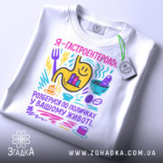 Футболка гастроентеролог, білого кольору, зображена з верхнього ракурсу, фон світло-сірий, принт кольоровий з жовтим шлунком, смайликами та предметами, що символізують гастрономію, з етикеткою на комірці.