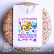 Футболка гастроентеролог, білого кольору, розкладена на плетеній кошику, фон світло-сірий, принт кольоровий з жовтим шлунком, смайликами та предметами, що символізують гастрономію.