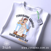 Футболка геніальний адвокат, біла, складена на столі, фон світло-сірий, принт з адвокатом у білому одязі, що тримає ваги та меч, текст чорний і жовтий, на комірці видно етикетку.