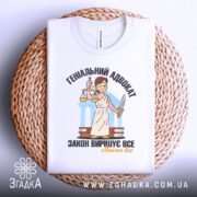 Футболка геніальний адвокат, біла, складена на плетеному кошику, фон світло-сірий, принт з адвокатом у білому одязі, що тримає ваги та меч, текст чорний і жовтий, видно етикетку на комірці.