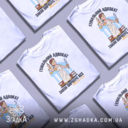 Футболка геніальний адвокат, біла, зображена на манекені з блискучого металу, фон світло-сірий, принт з адвокатом у білому одязі, що тримає ваги та меч, текст чорний і жовтий.
