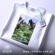 Футболка геть з України, біла, складена на поверхні, фон світлий, принт зображує жінку та істоту на городі, текст на принті: 'Геть з України, москаль некрасивий!', з етикеткою на комірці.