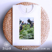 Футболка геть з України, біла, складена на кошику, фон натуральний, принт зображує жінку та істоту на городі, текст на принті: 'Геть з України, москаль некрасивий!'