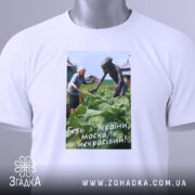 Футболка геть з України, біла, складена на поверхні, фон світлий, принт зображує жінку та істоту на городі, текст на принті: 'Геть з України, москаль некрасивий!', з етикеткою на комірці.