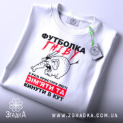 Футболка гніву білого кольору, зображена на столі, ракурс зверху, фон світлий, принт з малюнком гніву слона та текстом червоного і чорного кольору, на футболці є етикетка.